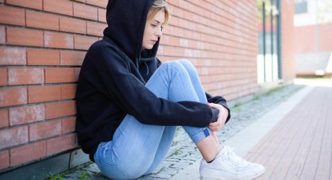 Young woman in a hooded sweatshirt sitting on ground with knees in her arms, looking down.