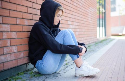 Young woman in a hooded sweatshirt sitting on ground with knees in her arms, looking down.