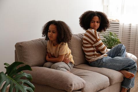Two young sisters sitting on a couch with their backs to each other, both looking angry.
