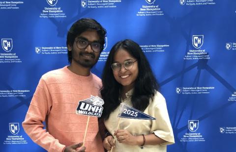 Two students with dark hair and glasses holding UNH wildcats and Class of 2025 photo props in front of blue background