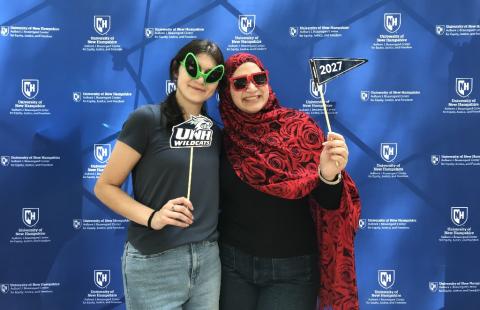 two students wearing green and red silly glasses holding UNH wildcats photo props in front of blue background