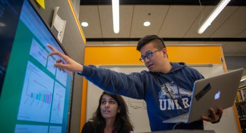 UNH Student pointing at screen with charts on it holding a laptop