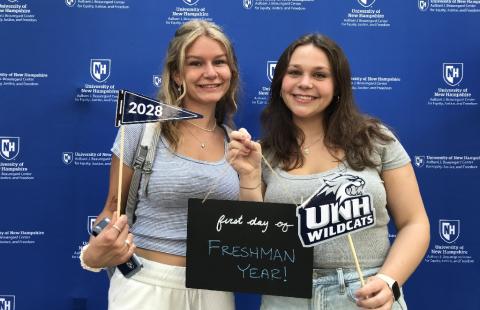 Two first year students holding a sign that says "First day of Freshman Year" with UNH wildcats and class of 2028 photo props
