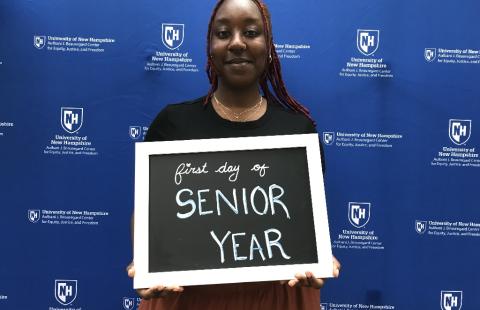 Student with dark red hair holding a sign that says, "First day of Senior Year" in front of blue background