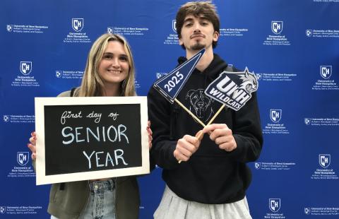 Two students holding sign that says "First day of senior year" in front of blue photo background