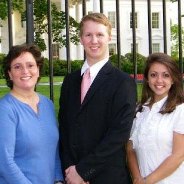 A man and two women stand outside a fence around a white building, all dressed in business attire