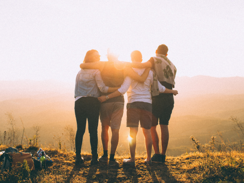 Four people standing with arms around each other's shoulders and backs to camera facing sunset 