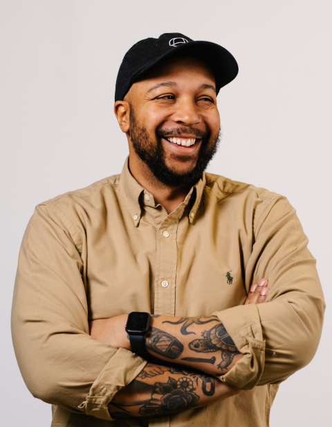 A man with a beard smiling wearing a black hat and a tan shirt