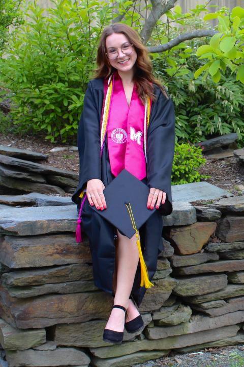 Photo of Katherine in graduation regalia sitting on a stone wall
