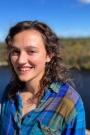 Val, a woman with brown curly hair, wears a blue flannel shirt and stands in front of a wooded pond backgorund.