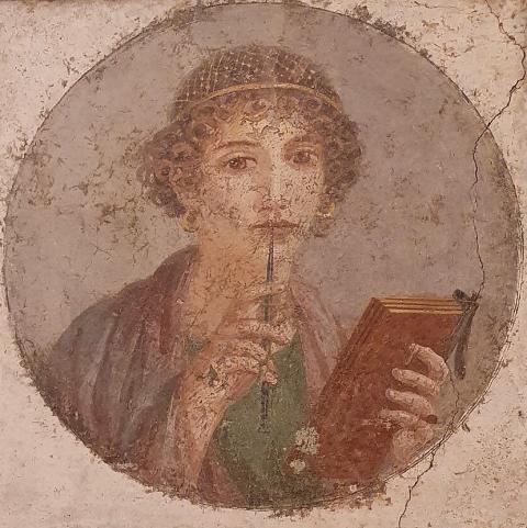 A picture of a fresco - painting on a cracked wall, of a woman in blue and green robes with curly hair, holding a book in one hand and a writing utensil in another. 