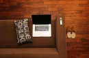 picture of a silver laptop sitting on a brown couch with a remote control on the arm of the couch, a pillow with letters next to it, and a pair of sheepskin slippers on a wood floor