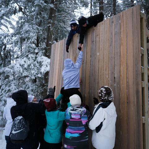 students assisting another student over a wooden wall 