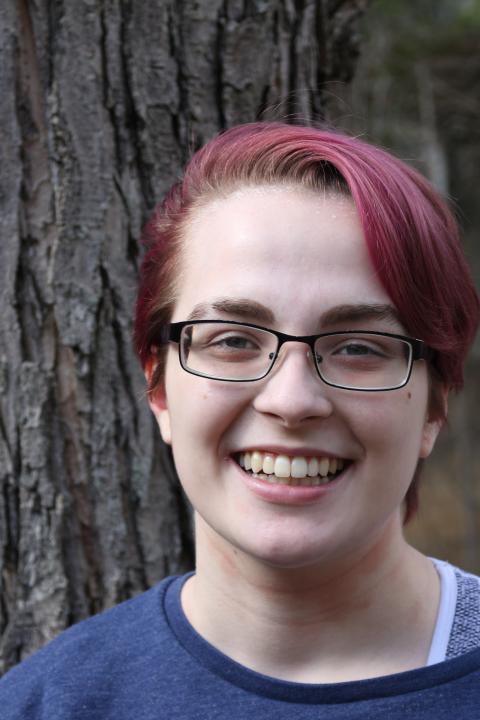 Young man with purple hair and glasses smiles in front of a tree