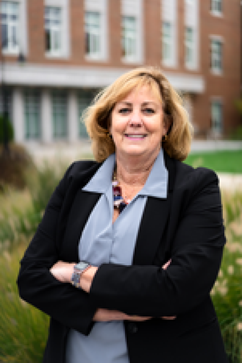 Headshot of Dean Lucy Gilson; wearing a dark blazer and light blue shirt standing with arms crossed in front of a brick building