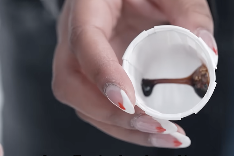 Close up photo of a woman's hand holding a small white cup featuring a dark syrup sample inside