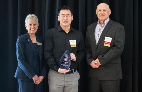 A man holding an award stands between a woman on the left and a man on the right