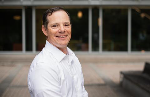 Portrait of Peter Zaimes wearing a white button down shirt and standing in front of an academic building