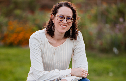Portrait of a woman in glasses wearing a white sweater, sitting atop a picnic table in front of a wooded backdrop