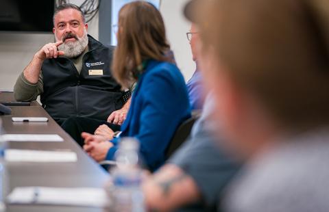 A man with a white beard wearing a black vest has a conversation with several people around a conference room table