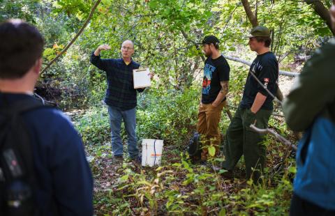 A man wearing a flannel shirt and glasses and holding a clipboard speaks to a group of students gathered around him in the woods