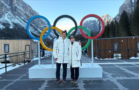A man and woman in matching white puffer jackets stand in front of the Olympic rings and snow-capped mountains