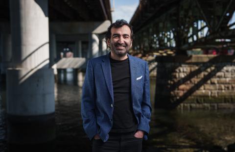 Man with dark hair, beard, and blue sportcoat stands under a highway bridge