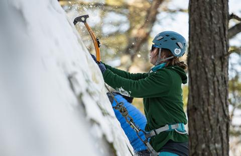 A woman in a helmet and green sweatshirt drives a climbing tool into an icy cliff face as she climbs up
