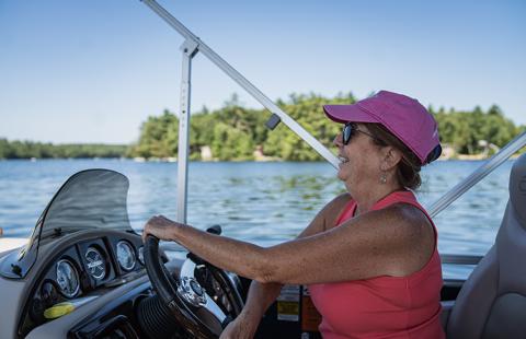 A woman wearing sunglasses steers a boat with a view of the lake and a small island featuring green trees in the background