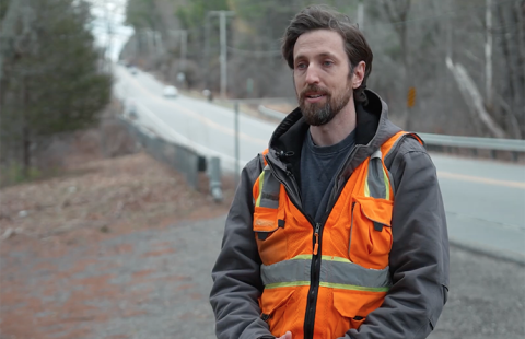 Screenshot from a video of a UNH researcher in an orange safety vest talking alongside a roadway