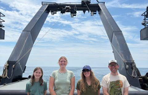 Three female students and one male researcher stand on board a research vessel.