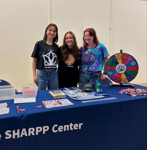 Three students stand at a SHARPP Center table with informational materials and a colorful prize wheel