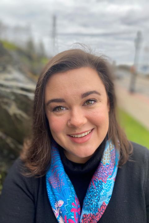 A photograph of Caroline. She wears a black shirt and colorful blue scarf. She is smiling and looking straight ahead.