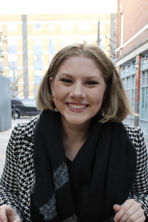 Headshot of Madison Wildey. She is looking directly into the camera and smiling. She wears a black and white jacket and a black scarf.