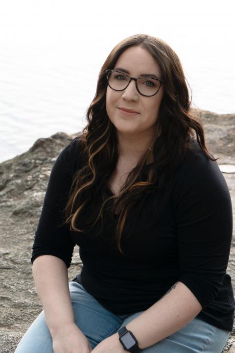 Julia sits facing the camera. She wears a black shirt and glasses and is smiling.