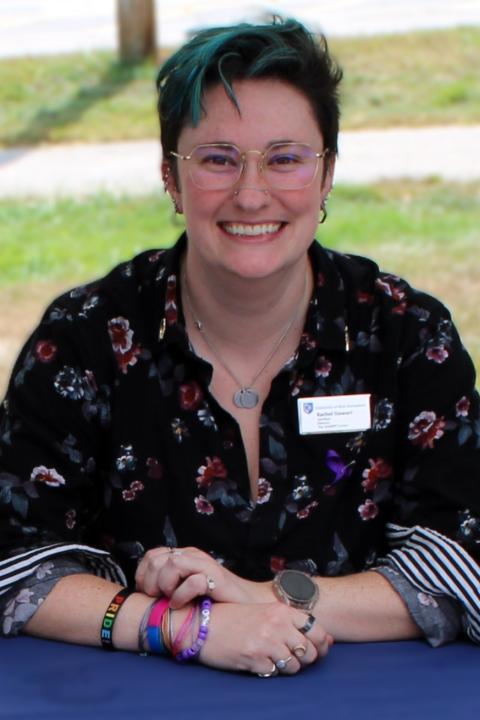 A photo of Rachel, a white nonbinary person. They have short, dark hair that is highlighted teal. They are sitting behind a table with their arms crossed over a blue tablecloth. 