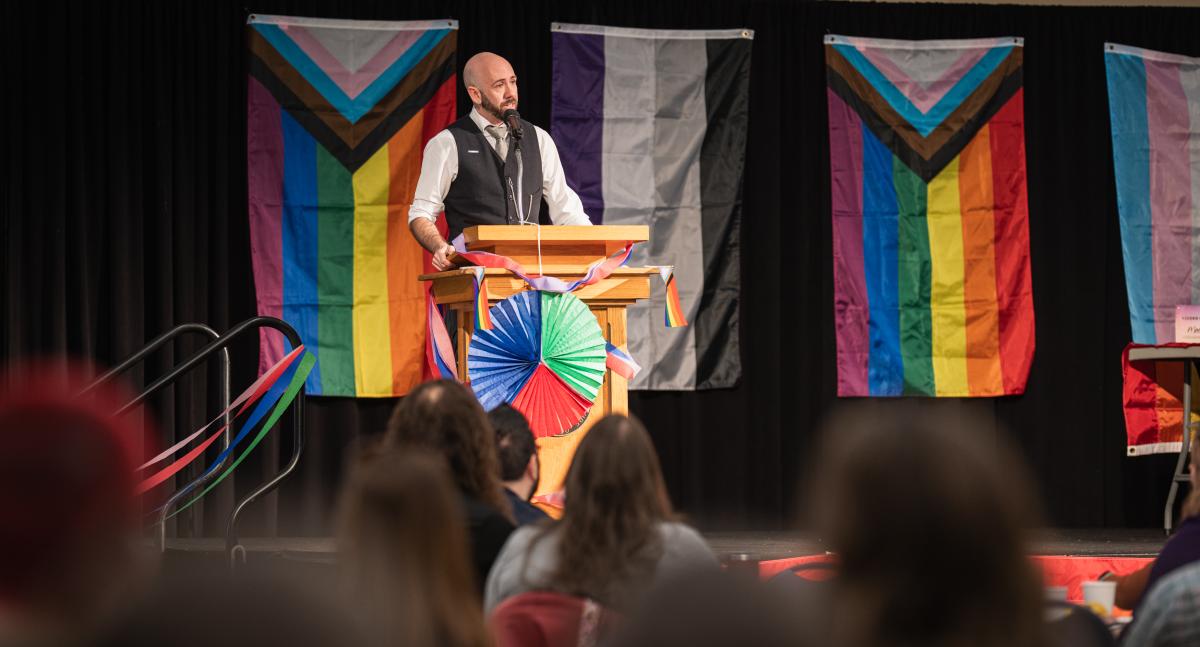 Mason Dunn, keynote speaker at Pride and Pancakes, stands at a podium in front of flags representing the LGBTQIA+ community during the Pride and Pancakes breakfast