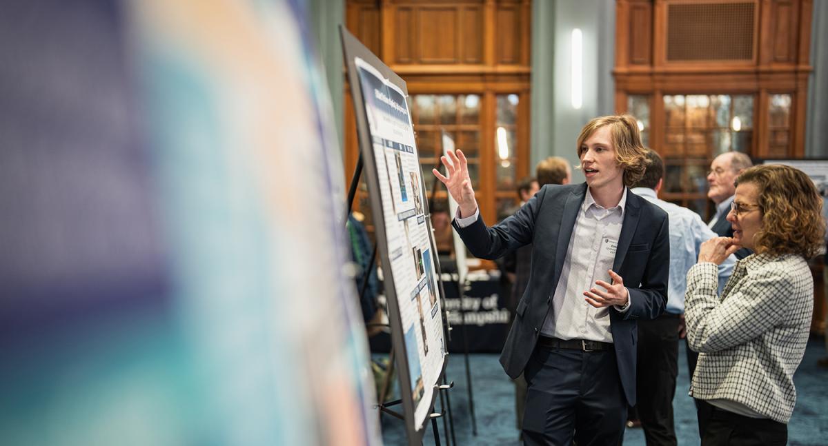 A male student in a suit coat gestures toward a poster while speaking to a woman