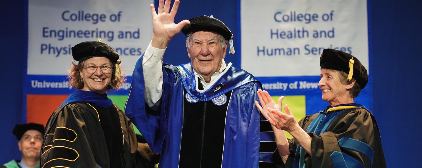 A man waves from the stage as he receives an honorary stole