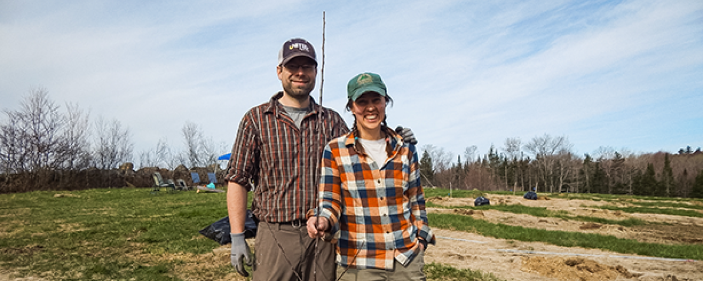A man and woman, both wearing flannel shirts and baseball caps, stand together and smile next to a freshly-planted sapling.
