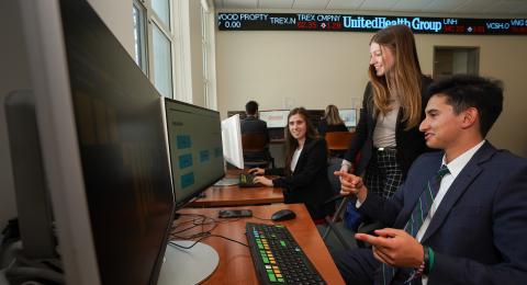 Two professionally dressed students sit at computers in a naturally lit room. A third student stands between, as they work together.