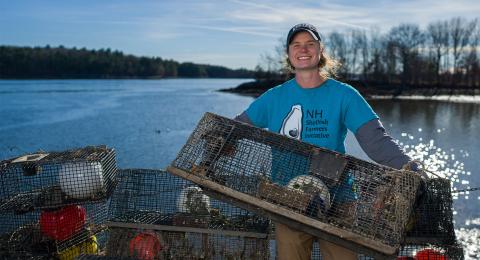 UNH COLSA alum Kelsey Meyer stands on a boat holding up a shellfish trap and wearing a t-shirt with the words NH Shellfish Farmers Initiative on it. Kelsey is white with hair tucked beneath a ballcap and the t-shirt is teal.