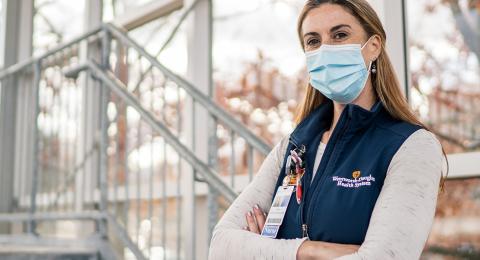 Nurse wearing a surgical mask standing in a glass-walled stairwell with a view of trees with their arms folded