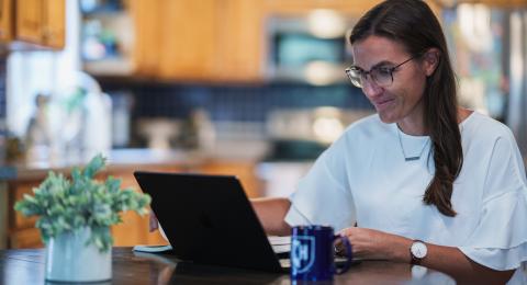 A woman with dark hair wearing glasses works at a computer on a circular table with a kitchen in the background