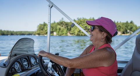 A woman wearing sunglasses steers a boat with a view of the lake and a small island featuring green trees in the background