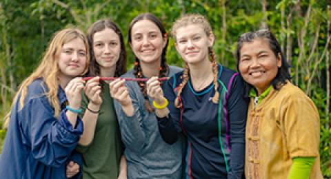 five people standing side by side outside smiling and some are holding a red stick
