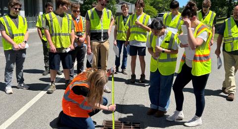 Students wearing high-viz vests stand watching an instructor measure a storm drain