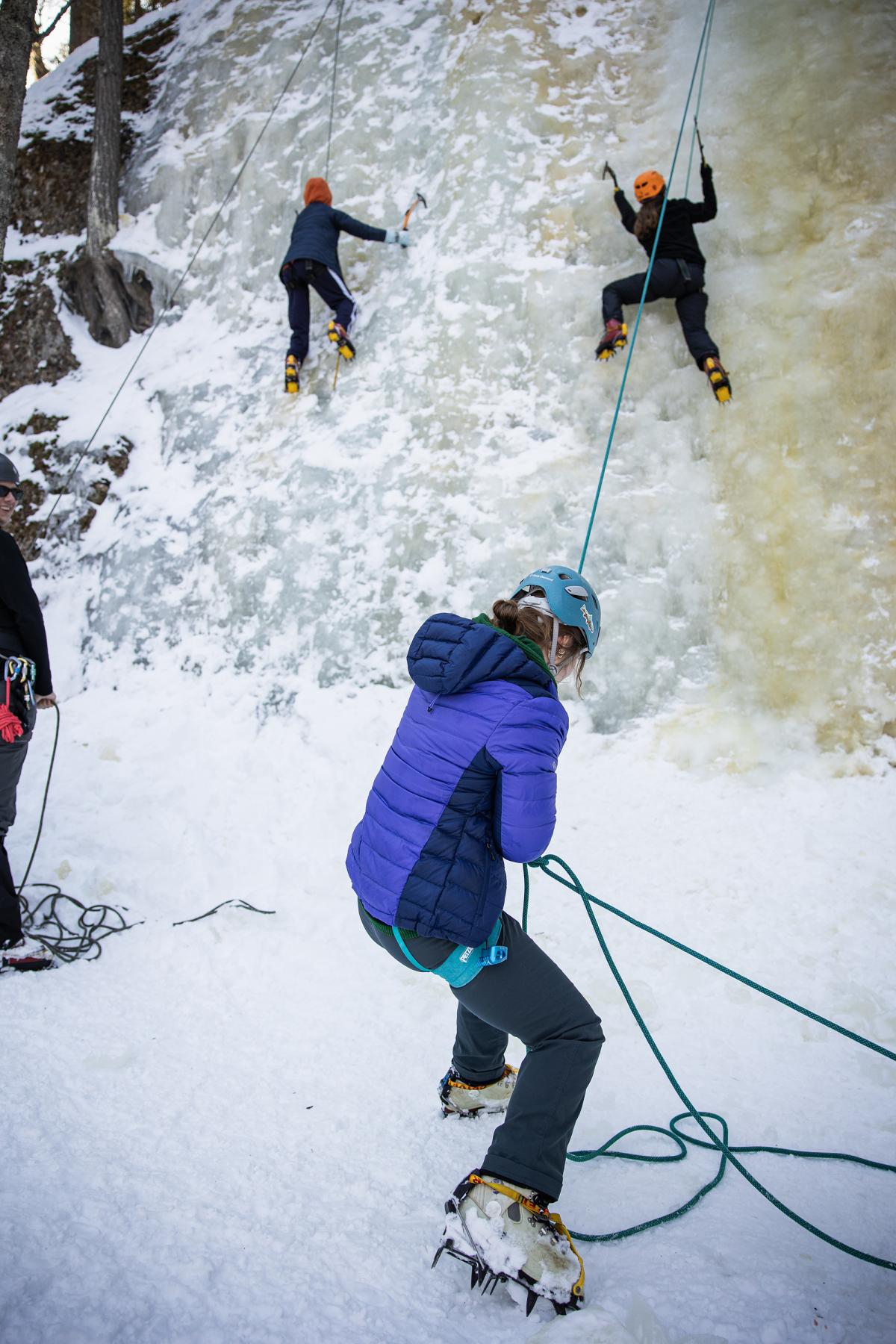 A student in a blue jacket belays from ground level while two students climb an icy rock face