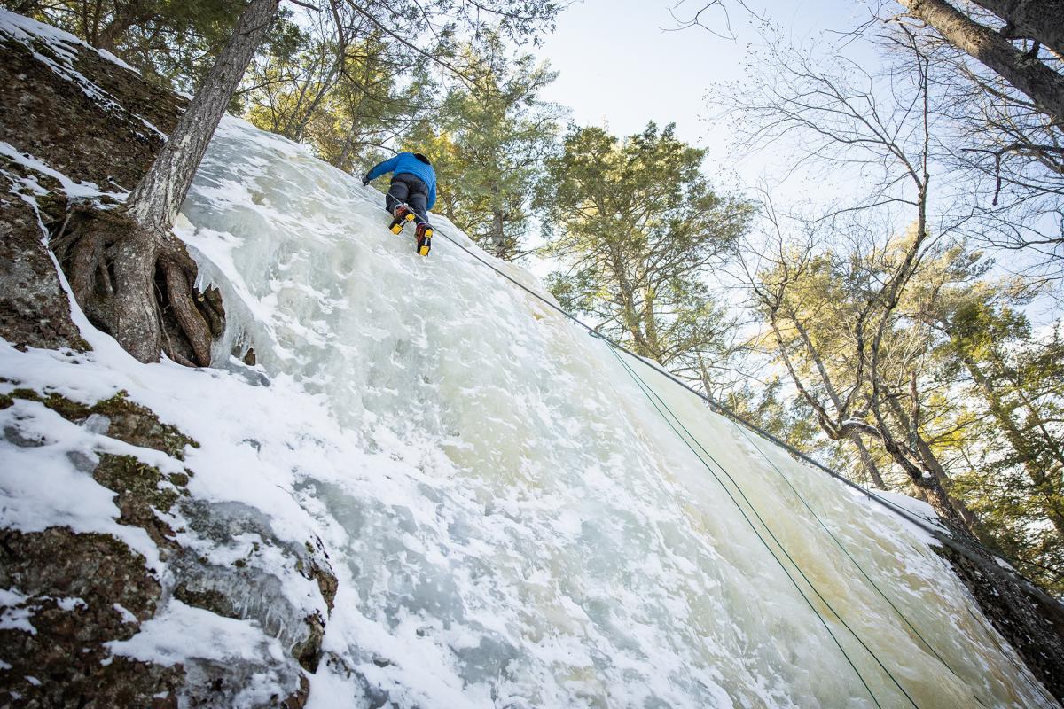 A student is visible at the very top of an icy cliff after ice climbing the surface