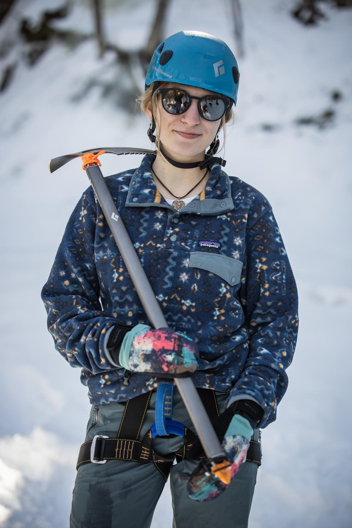 A woman in a blue helmet and sunglasses holds an ice climbing tool over her right shoulder